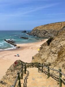 Idyllic rocky beach with crystal clear waters, sandy shore, and cliffside steps under a clear blue sky.