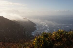 Breathtaking sunrise view of Fisterra's rugged coast with misty waves and distant lighthouse.