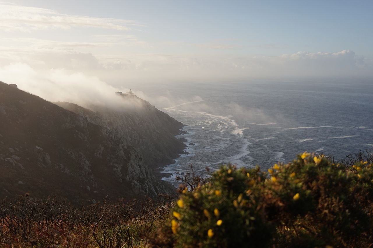 Breathtaking sunrise view of Fisterra's rugged coast with misty waves and distant lighthouse.